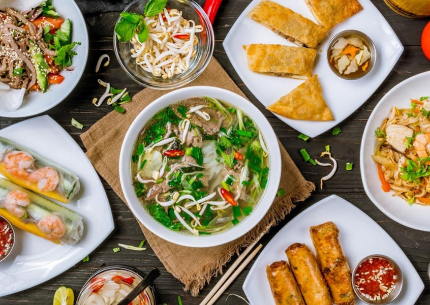 Vietnamese cuisine spread featuring pho soup, fresh spring rolls, fried rolls, and various side dishes on a wooden table