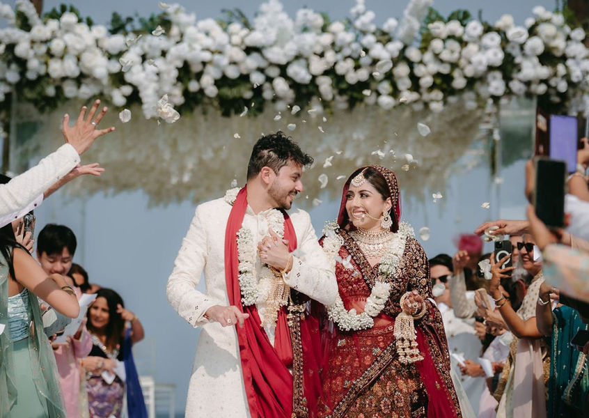 Bride and groom walk under white floral arch amid petal shower