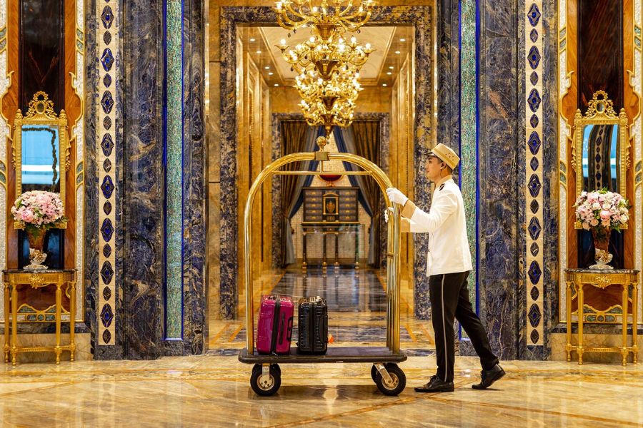 The Reverie Saigon luxury lobby with bellhop pushing a luggage cart past ornate marble and mosaic pillars and a crystal chandelier